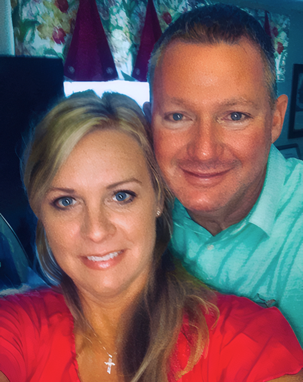 Man and Woman named Andy and Mac smiling looking at the camera. Their heads are touching, Mac is in the front and is wearing an orange shirt and has her hair in a pontytail. Andy is behind her and he has short hair and is wearing a light blue shirt